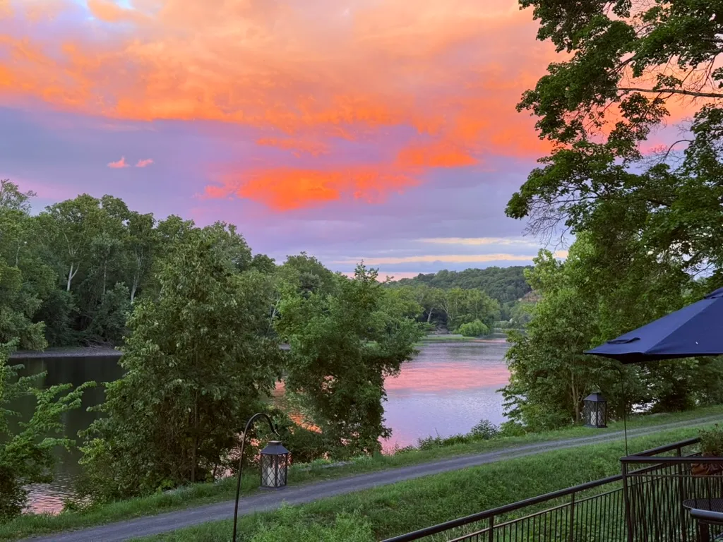 A wide river at sunset under orange and lavender clouds, framed by trees, with lanterns along a path in the foreground.